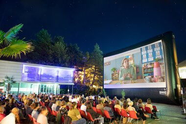 In einem Freiluftkino in der Stadt Bludenz sitzen Zuschauerinnen und Zuschauer und schauen einen Film auf einer großen Leinwand. Es ist bereits dunkel. | © Anna Salcher