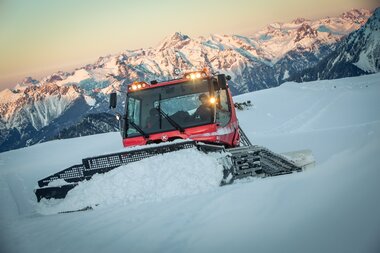 Ein PistenBully schiebt im Vordergrund den Schnee. Im Hintergrund färben sich die Bergspitzen bei Sonnenuntergang rosa. | © Bergbahnen Brandnertal, Michael Marte