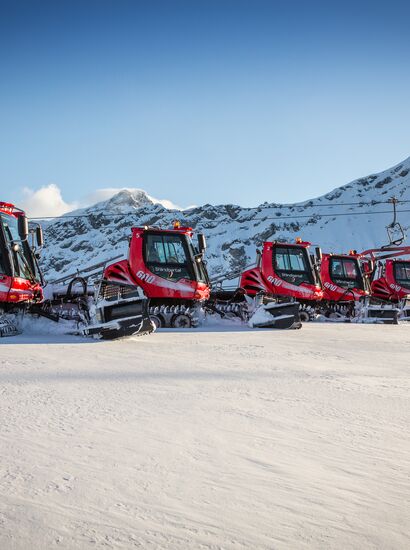 Acht PistenBullys stehen im Skigebiet Brandnertal in einer Reihe. Im Hintergrund, ein Bergpanorama. | © Bergbahnen Brandnertal, Michael Marte