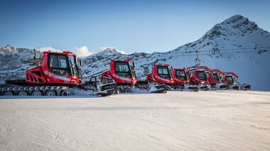 Acht PistenBullys stehen im Skigebiet Brandnertal in einer Reihe. Im Hintergrund, ein Bergpanorama. | © Bergbahnen Brandnertal, Michael Marte